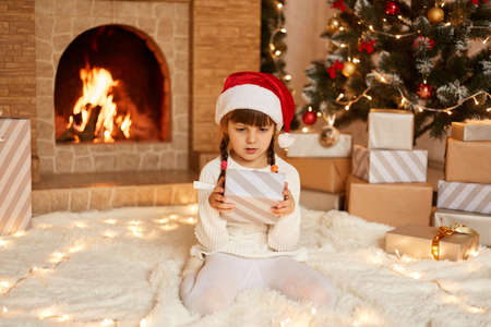 Cute little female kid wearing white sweater and santa claus hat, posing in festive room with fireplace and Xmas tree, holding gift box in hands, looking at present with astonishment.の写真素材