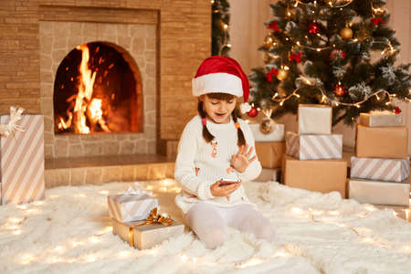 Happy positive little girl wearing white sweater and santa claus hat, sitting on floor near Christmas tree, present boxes and fireplace, having video call with friends via smartphone.の写真素材