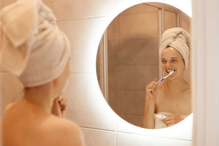 Portrait of optimistic winsome woman brushing her teeth, having hygiene procedures after taking shower in bathroom, standing with white towel on her hair.の写真素材