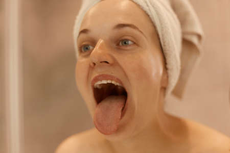 Close up portrait of young adult beautiful woman posing in bathroom, showing her tongue, being wrapped in white towel, doing hygienic procedures at home in morning.の写真素材