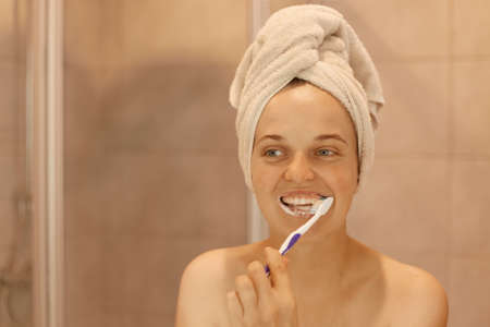 Smiling happy attractive woman in good mood brushing her teeth, having hygiene and beauty procedures at home in bathroom, being wrapped in white towel.の写真素材