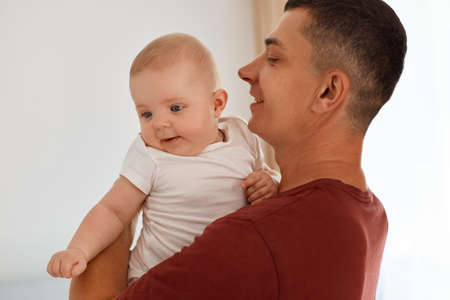 Indoor shot of handsome father wearing burgundy t shirt with charming infant daughter, family expressing happiness and positive emotions, parenthood and childhood.の写真素材