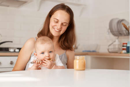 Portrait of smiling happy woman sitting at table in light room with her infant daughter, looking at...