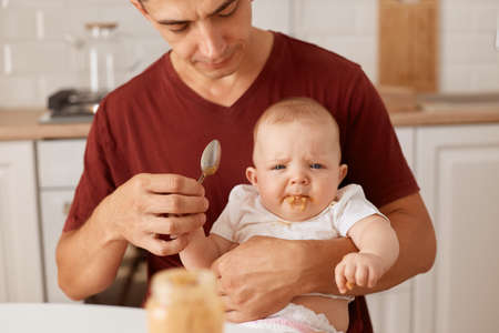 Concentrated serious brunette man feeds little daughter while sitting at table in kitchen, father feeding up kid with fruit or vegetable puree, parenthood.の写真素材
