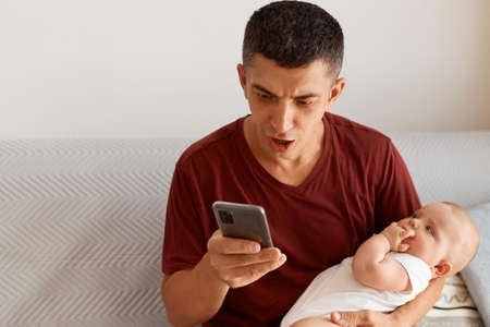 Shocked emotional man with dark hair holding baby girl in hands, using cell phone, looking at device display with scared angry facial expression, reading message with bad news.の写真素材
