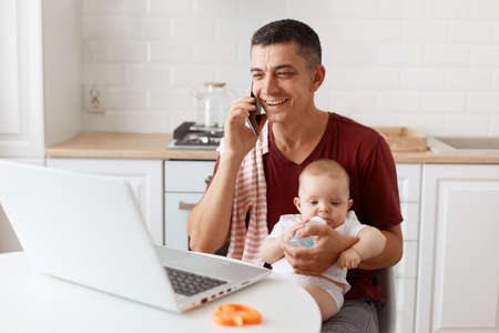 Smiling happy man wearing burgundy casual t shirt with towel on his shoulder, looking after baby and...