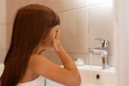 Indoor shot of dark haired female child standing in bathroom and washing her face after waking up or before going to bed, hygienic procedures at home.の写真素材