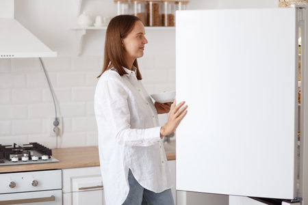Profile portrait of beautiful young adult woman wearing white shirt, looking smiling inside fridge with pleasant smile, holding plate in hands, posing with kitchen set on background.の写真素材