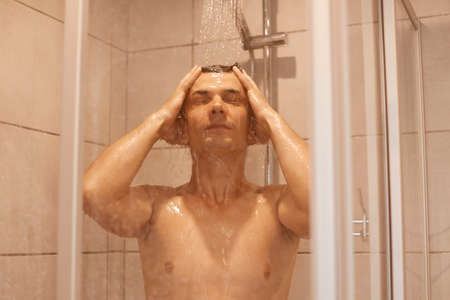 Portrait of young adult man washing his body skin and hair under a shower at home, bathroom interior. Healthy handsome man lifestyle, well being and grooming.の写真素材