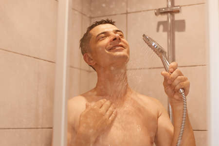 Indoor shot of satisfied positive brunette handsome man, taking shower, posing naked under water, expressing positive emotions, relaxing before going to bed.の写真素材