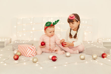 Portrait of little sisters wearing casual clothing and party hoops sitting on bed in light festive room playing with baubles for x mas tree decorations, spending new year together at home.の写真素材