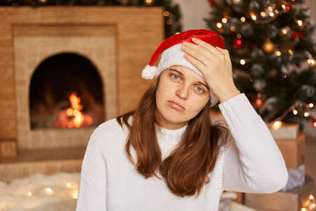 Unhealthy Caucasian woman wearing white sweater and santa claus hat, sitting near fireplace and xmas tree and keeping hand on her forehead, suffering headache, flu symptoms.の写真素材