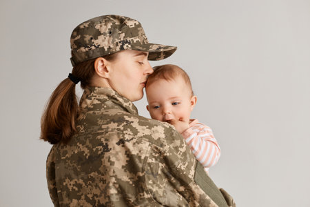 Back side portrait of young adult woman soldier wearing camouflage uniform, returning home, embracing her little girl daughter and kissing her head, expressing love.の写真素材