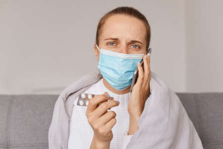Indoor shot of unhealthy woman wearing white sweater and surgical mask, looking at camera, holding...