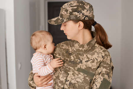 Indoor shot of young adult woman soldier wearing camouflage uniform, returning home after served in...