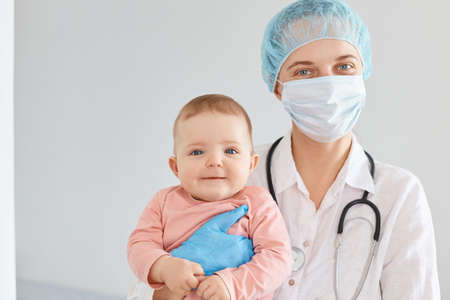Indoor shot of woman family doctor wearing medical uniform and surgical mask holding infant baby in hands, looking at camera, examines children health.の写真素材