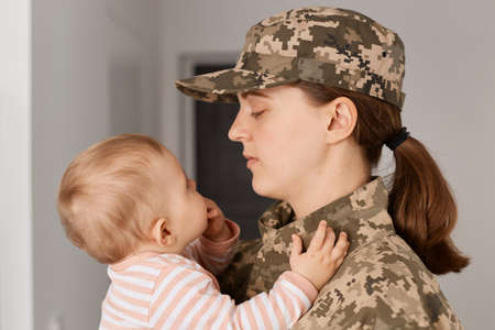 Side view of portrait of lovely young adult woman soldier wearing camouflage uniform, returning home and meets with her little daughter, looking at daughter, cant believe their meet.の写真素材