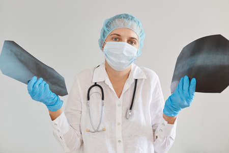 Indoor shot of confused female doctor wearing medical uniform, cap and gloves, holding two x rays,...