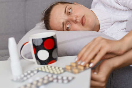 Side view portrait of sick woman lying in bed, expressing sadness and sorrow, feeling bad, taking pills from the table to treat grippe or influenza symptoms.の写真素材