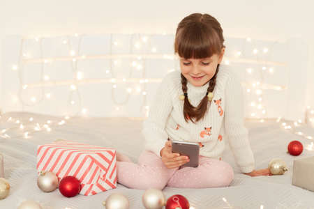Smiling dark haired female with pigtails wearing white sweater using mobile phone, watching cartoons or playing games while sitting on bed with Christmas decorations.の写真素材