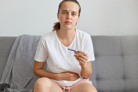 Indoor shot of sad young adult Caucasian woman holding pregnancy test in hands, touching her belly, looking at camera with sorrow, sitting on gray sofa at home in living room.の写真素材