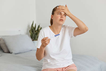 Sad unhappy young adult pregnant woman with pregnancy test posing in her bedroom, wearing white t...