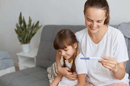 Caucasian young adult woman wearing white t shirt holding pregnancy test in hands, sitting on sofa...