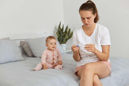 Portrait of sad frustrated young adult pregnant woman with pregnancy test posing in her bedroom with toddler baby, wearing white t shirt, being sad to see positive result.の写真素材