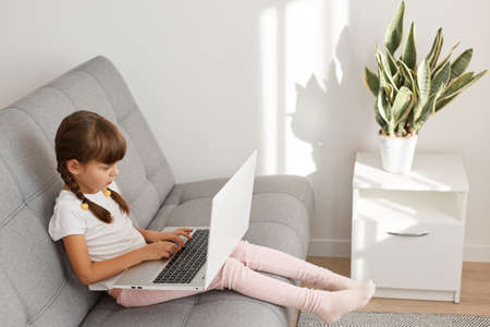 Profile indoor shot of little cute dark haired female child wearing white casual t shirt using...