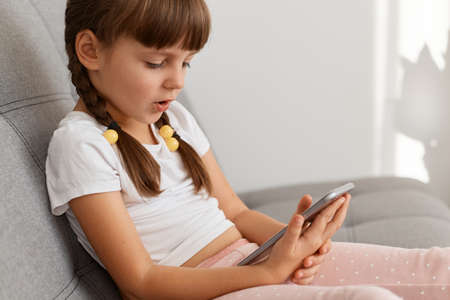 Side view portrait of calm little girl wearing white t shirt sitting on sofa at home, using mobile...