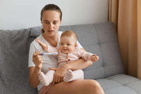 Shocked surprised woman with pigtail wearing white t shirt sitting on sofa with her little daughter...