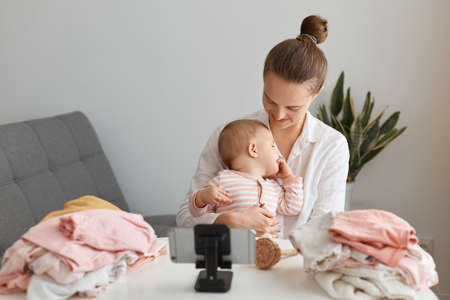 Indoor shot of young adult woman blogger wearing casual clothing and her kid sitting in front of phone on tripod and shooting video, being surrounded with baby clothing, boasting her purchases to followers.の写真素材