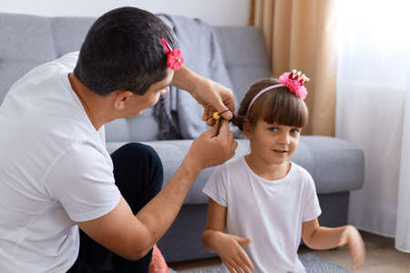 Indoor shot of happy family sitting on floor at home in living room, father braiding girl's hair, family time at weekend, happy child with hie lovely daddy.の写真素材
