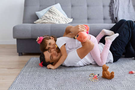 Indoor shot of cute female child hugging happy dad lying on floor while spending time together, family with hairpins expressing positive emotions and laughing out loud.の写真素材