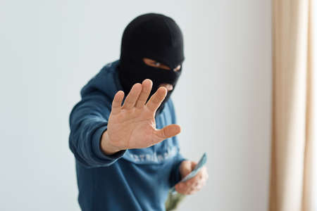 Portrait of man robber wearing blue hoodie and black balaclava, looking at camera, standing with outstretching arm, showing stop ban gesture, holding money in hand.の写真素材