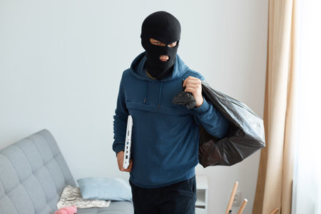 Indoor shot of serious male burglar wearing dark blue hoodie and black mask posing in the house he...