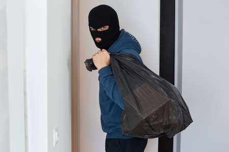 The thief holds a bag of stolen things in his hands, looks at the camera, is pleased that he found many expensive things to steal, male burglar wearing dark blue hoodie and black mask.の写真素材