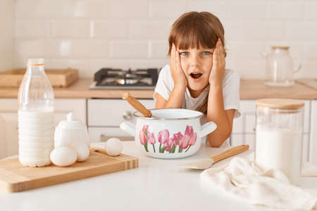 Astonished shocked charming little girl preparing dough for baking, posing with open mouth and covering cheeks with palms, looking at camera, does not know how to make dough.の写真素材
