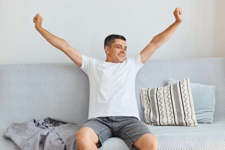 Indoor shot of happy positive attractive brunette man wearing white shirt and jeans, sitting on gray couch in light living room, stretching arms, just wake up in high spirit.の写真素材