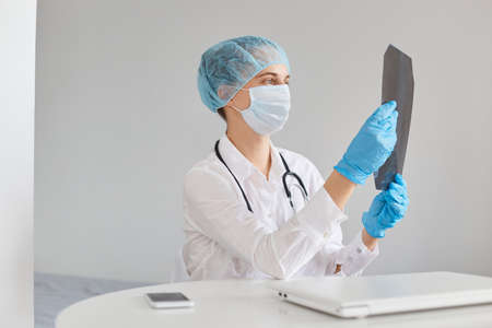 Concentrated female doctor wearing medical cap, surgical mask, rubber gloves and gown, sitting at table and holding x ray picture, examining scan of lungs of patient.の写真素材