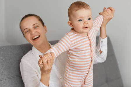 Portrait of Caucasian mother playing with her infant baby, family spending time together, woman with hair bun laughing happily while sitting with her toddler daughter on sofa.の写真素材
