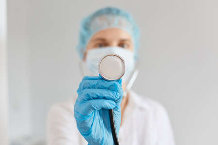 Indoor shot of Caucasian female doctor holding stethoscope in hand, standing indoor on white background, wearing gloves, medical mask ans gown, medicine and health care.の写真素材