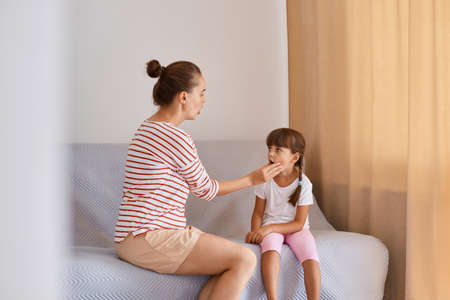 Horizontal shot of female speech therapist working with little girl, sitting on sofa near window, having language lesson for improving speaking, language therapist practicing sounds pronunciation.の写真素材