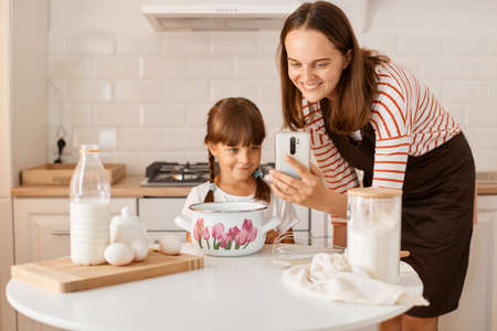 Portrait of smiling young adult woman cooking with her daughter in the kitchen, browsing internet for finding recipe of the pie or cake, family expressing positive happy expression.の写真素材