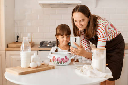 Indoor shot of Caucasian young adult woman cooking with her daughter in the kitchen, browsing internet for finding recipe, looking smiling at device, being happy to bake together.の写真素材