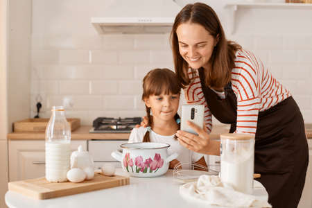 Young adult mother baking together with her daughter, sitting at table in the kitchen and finding...
