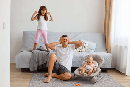 Portrait of tired exhausted handsome brunette father playing with his daughters while sitting on floor near sofa, wearing white t shirt and jeans short, male covering ears, because of noisy kids.の写真素材