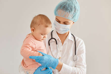 Image of charming baby and doctor pediatrician wearing medical cap, surgical mask, gown and rubber gloves, woman standing with toddler girl in her arms.の写真素材