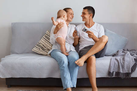 Smiling happy young family sitting on comfortable cough with their toddler daughter in living room,...