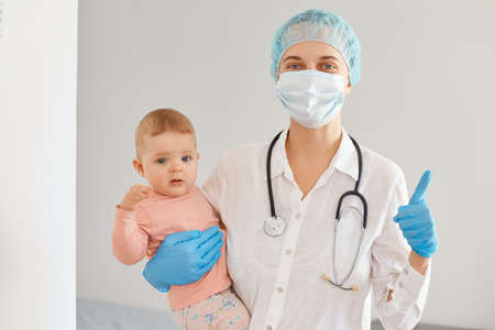 Image of positive young adult female pediatrist wearing medical uniform, holding infant baby girl,...